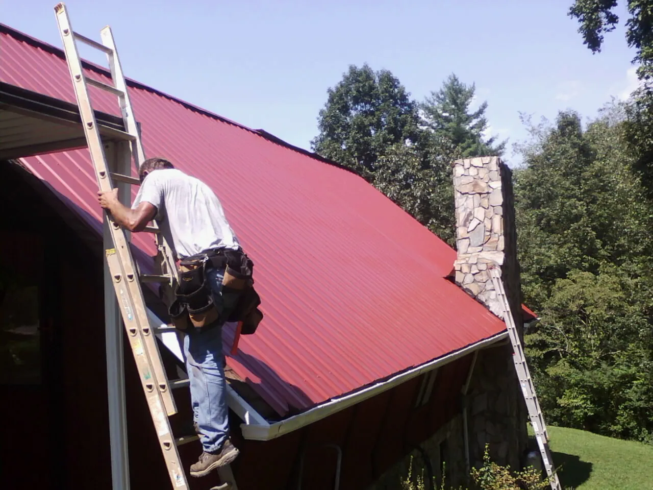 Roofer on a ladder beside a red metal roof and stone chimney — Soffit & Fascia Repair by Roofing Brothers in Leesville